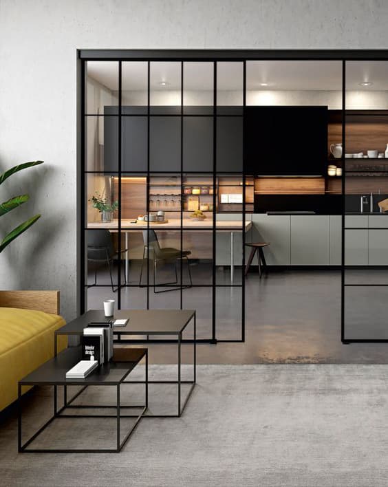 Minimalist black metal nesting coffee tables placed in front of a mustard yellow sofa, with a modern kitchen visible through glass grid sliding doors in the background.