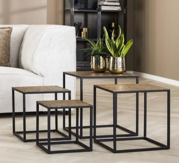 Set of four square nesting tables with textured wooden tops and sleek black metal frames, placed beside a cream-colored sofa and decorated with small indoor plants in metallic planters.