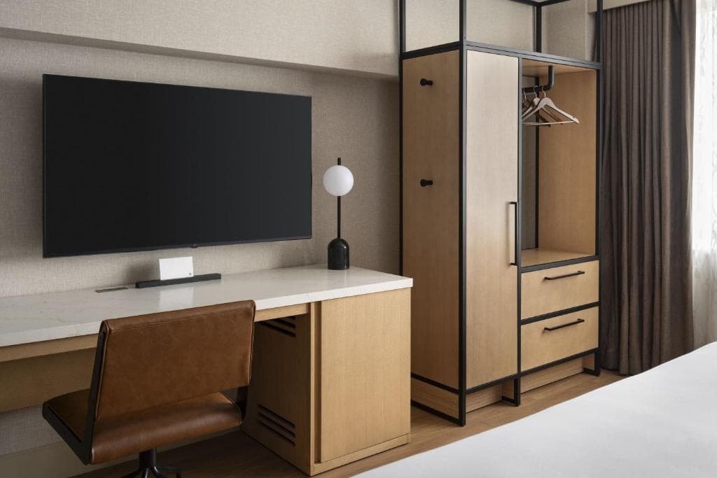 Modern hotel wardrobe with open hanging space, drawers, and a study desk beside a flat-screen TV.