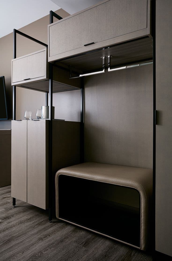 Contemporary hotel wardrobe unit with sleek wood finish, built-in bench, hanging rod, and upper storage cabinets.