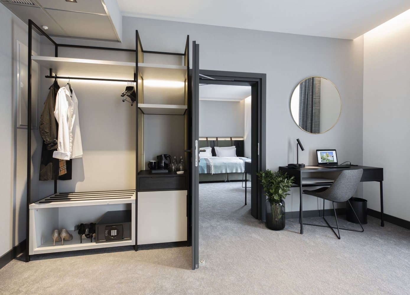 Open hotel wardrobe with hanging space, bench, minibar, and a modern work desk beside it.
