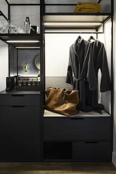 Dark-toned open wardrobe with robes, a brown bag, drawers, minibar setup, and glassware.