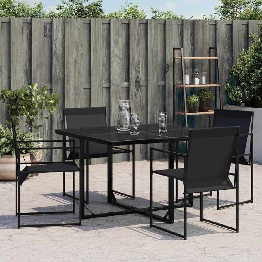 Black metal outdoor dining set with four chairs and a square table, placed on a stone patio with greenery in the background.