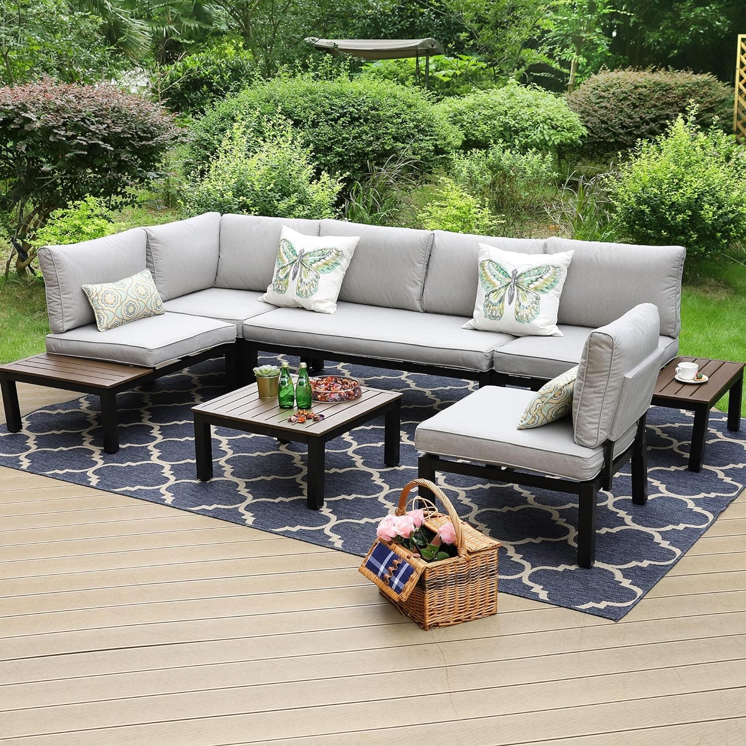 L-shaped outdoor sectional with gray cushions and butterfly pillows on a patterned rug in a garden setting.