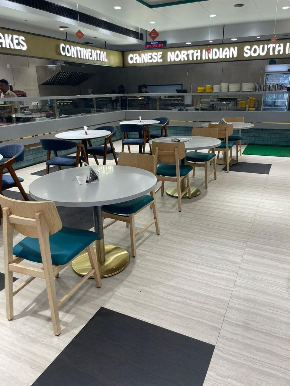 Modern food court with round tables, wooden chairs, and illuminated signs for various cuisines like Chinese and North Indian.