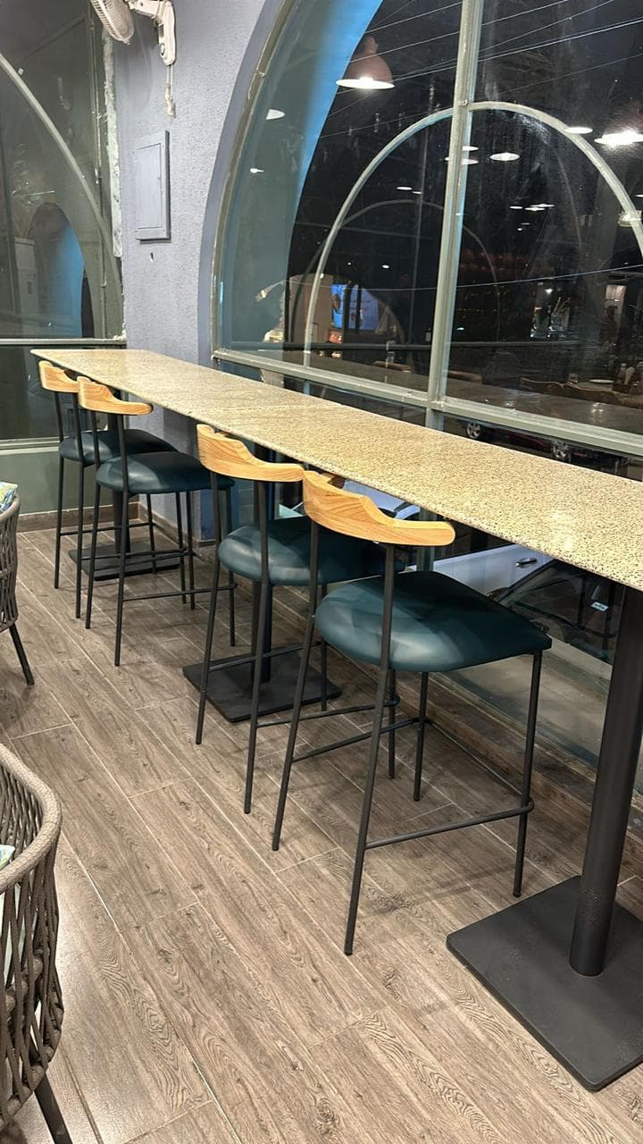High counter table with wooden barstools featuring black metal legs and cushioned seats, placed beside large arched windows.