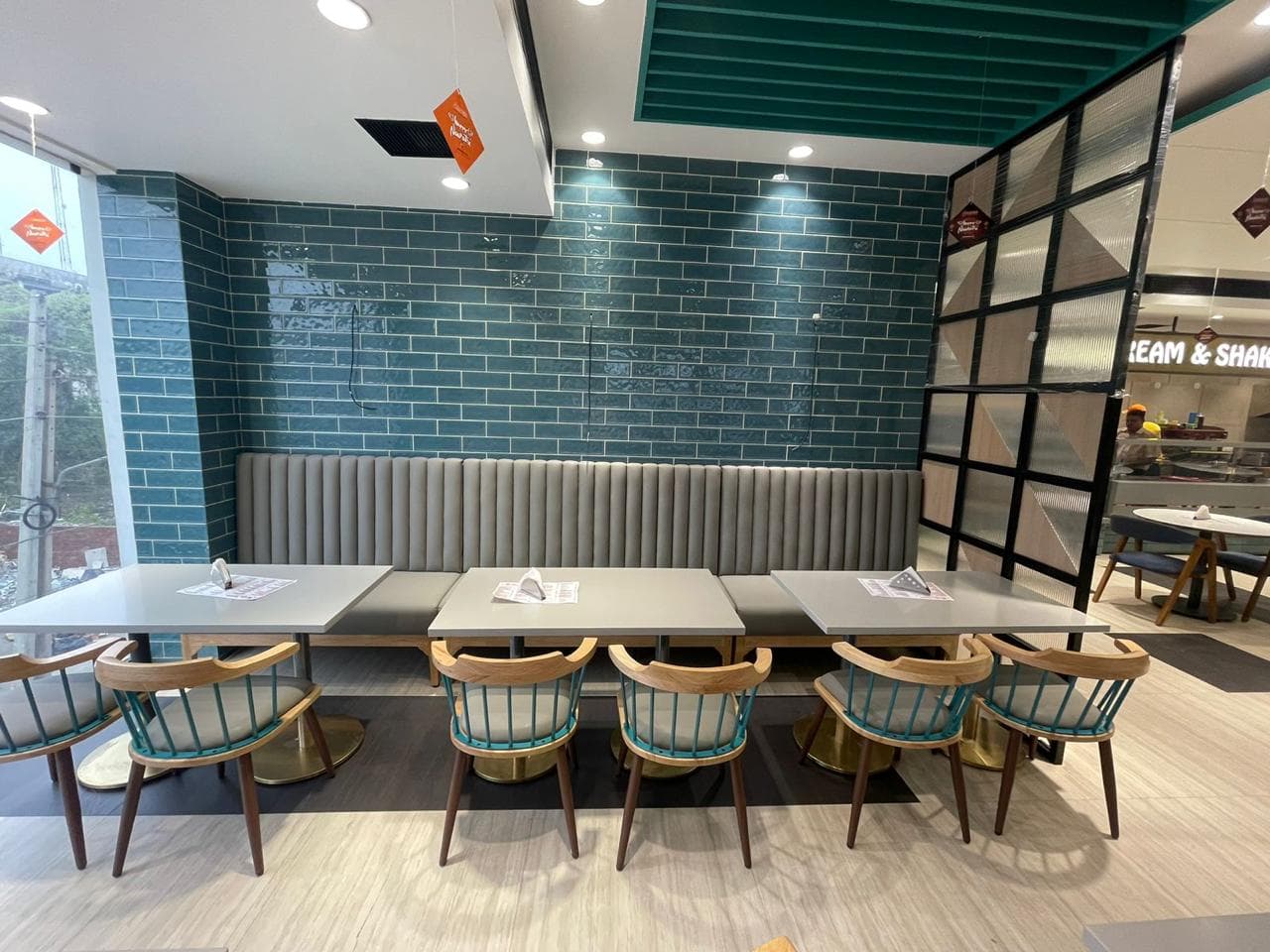 Restaurant seating with grey cushioned bench, teal brick wall, and wooden chairs with teal spindle backs.