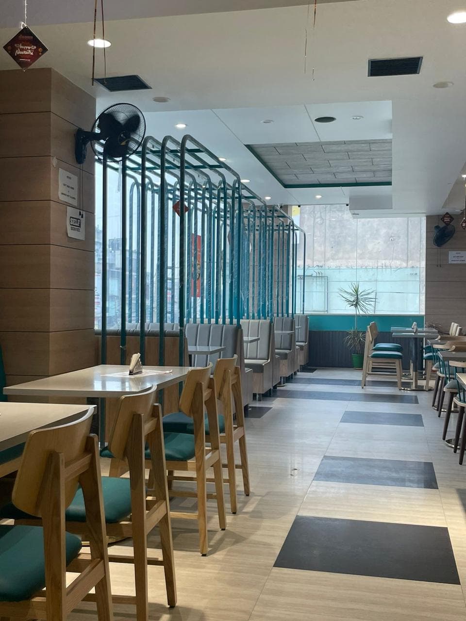 Modern restaurant interior with teal-accented wooden chairs, booth seating by large windows, and patterned tile flooring.