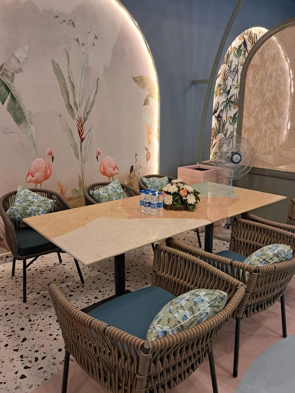 Elegant dining setup with rattan chairs, floral cushions, terrazzo flooring, and a flamingo mural backdrop in a cozy restaurant corner.