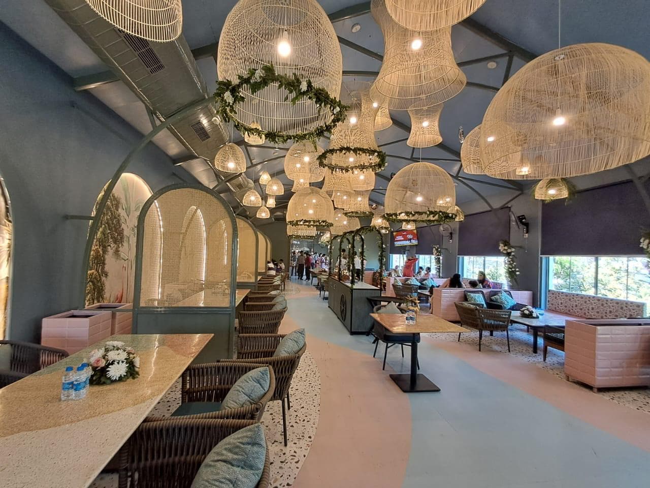 Spacious restaurant with wicker lighting, pastel seating, floral decor, and terrazzo flooring under a vaulted ceiling.
