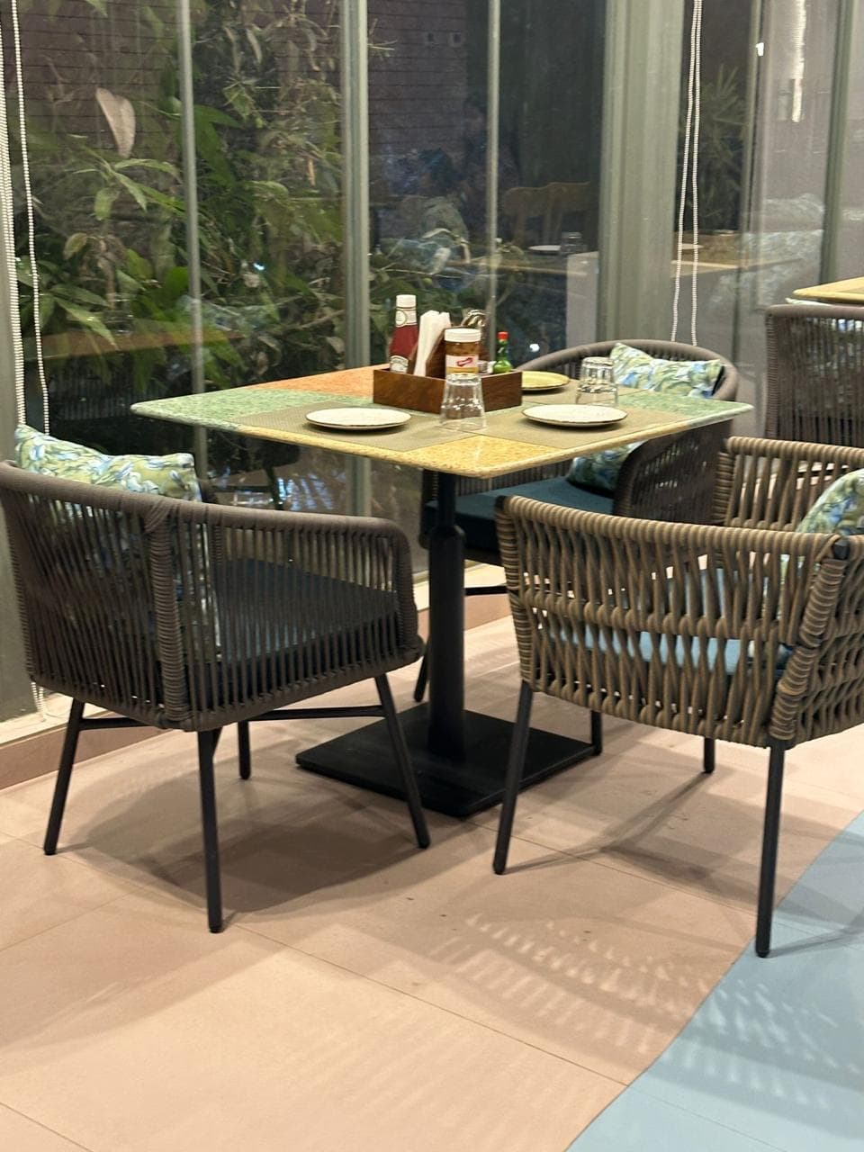 A cozy restaurant table for four with rope-woven chairs, floral cushions, and a colorful terrazzo-style tabletop near a glass wall.