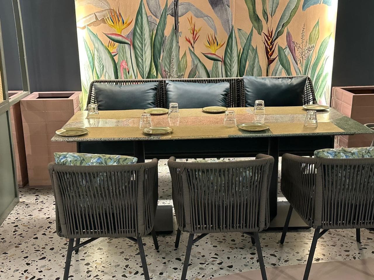 A six-seater restaurant setup with rope-woven chairs, a cushioned bench, and a vibrant tropical wall mural in the background.