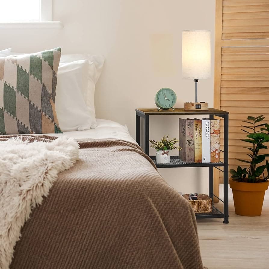 A cozy bedroom scene with a brown blanket, a checkered pillow, and a black metal bedside table holding books, a lamp, and a clock.