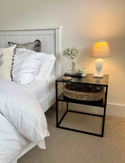 Black metal side table with a shelf, styled with a lamp, vase of flowers, and wicker basket, beside a white bed.