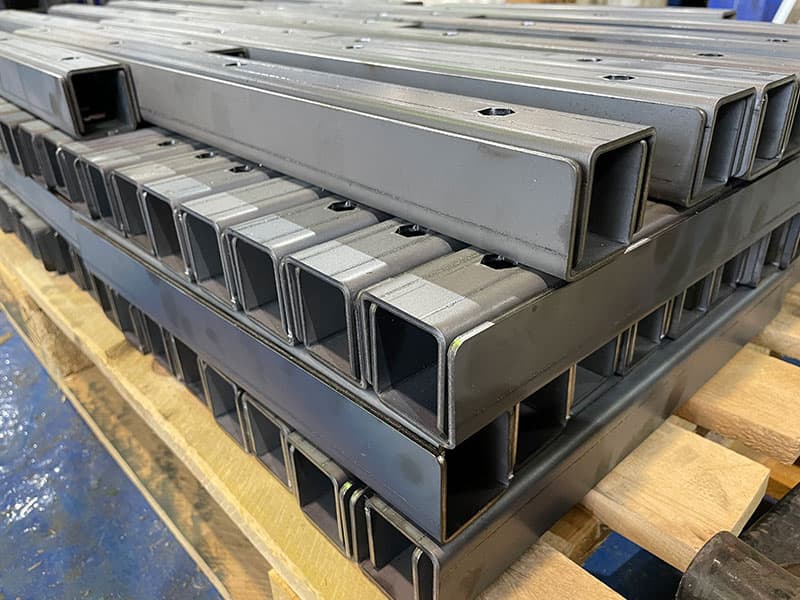 Stacked black metal rectangular tubing with pre-drilled holes, placed on wooden pallets for industrial use.