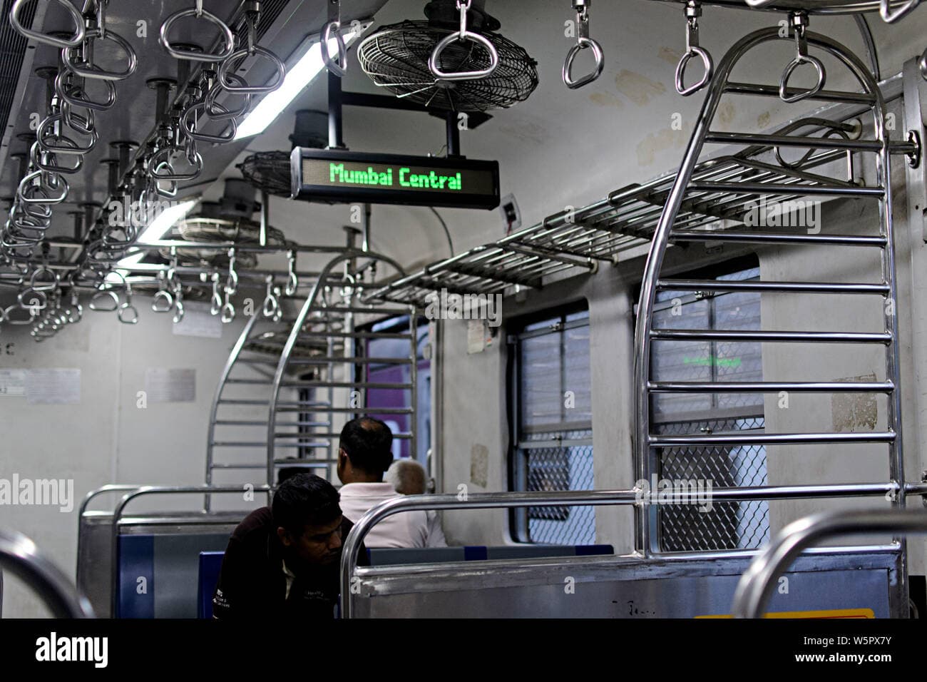 Interior of a Mumbai local train showing metal handrails, seating, luggage racks, and digital display reading 'Mumbai Central'.