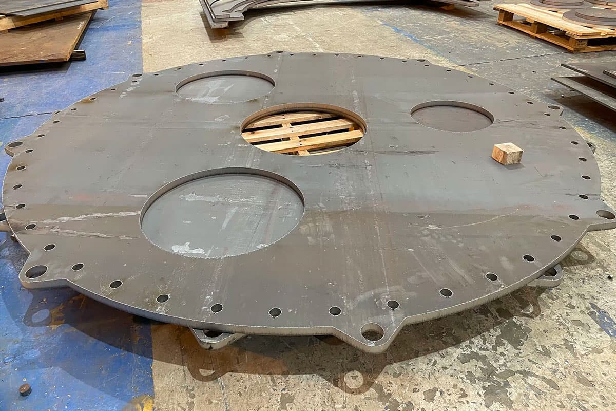 Heavy-duty circular steel base plate with multiple cutouts and bolt holes, placed on a workshop floor with pallets in the background.