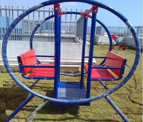 Circular outdoor swing set with two red seats facing each other, supported by a blue metal frame, placed on a grassy area.