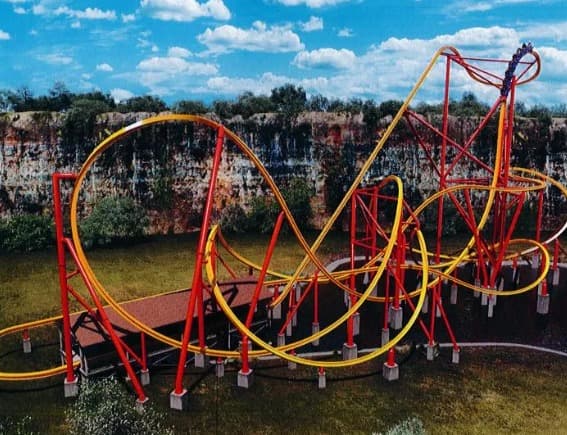 Large red and yellow roller coaster with multiple loops and twists set against a rocky cliff and blue sky.