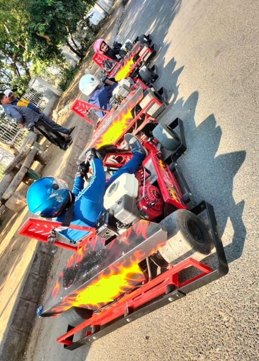 Three go-carts with vibrant flame designs parked on a sunny road, driven by riders wearing colorful helmets and gear, ready for action.