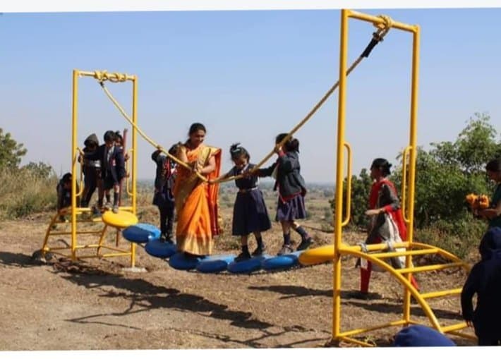 Children guided by a woman crossing a yellow rope bridge with blue balancing stones in an outdoor playground.
