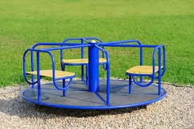 A blue metal merry-go-round with yellow seats on a gravel base, set in a grassy park.