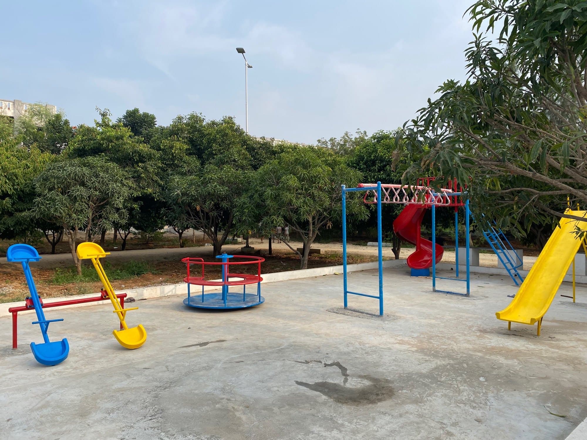 A playground with colorful equipment, including seesaws, a merry-go-round, slides, and monkey bars, surrounded by greenery.